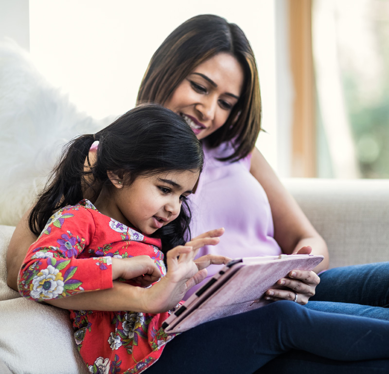 Woman and young daughter smile while using a tablet on the sofa.