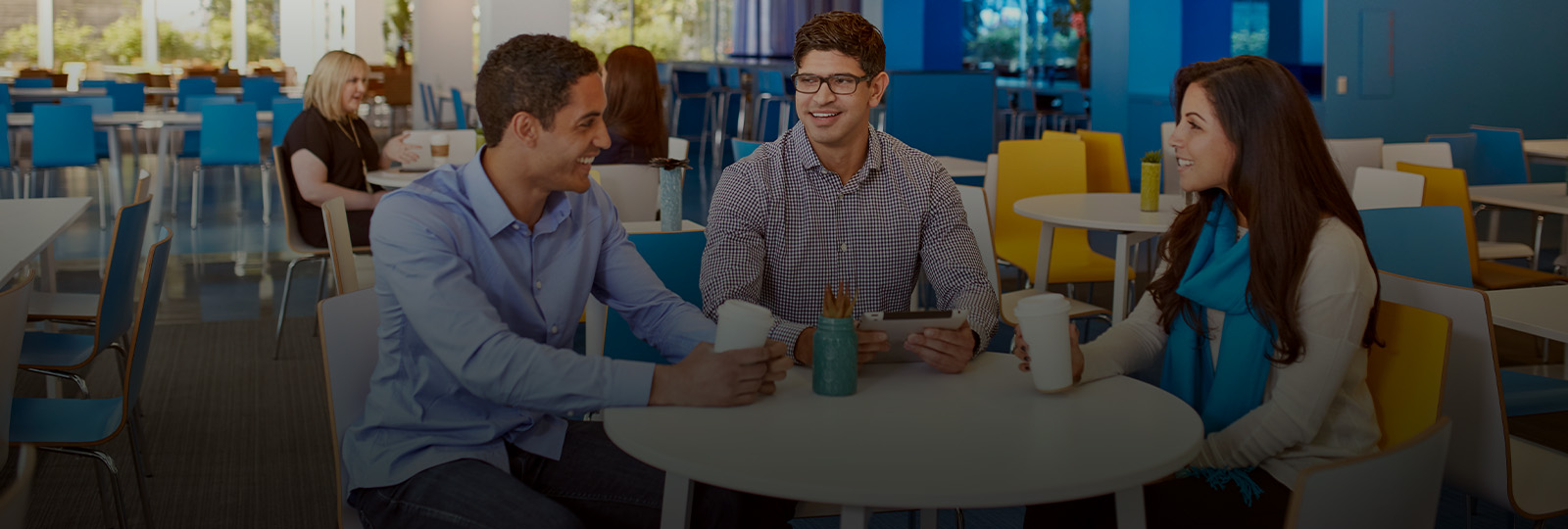 Three employees sitting at a table drinking coffee and using a tablet, with smiles on their face.