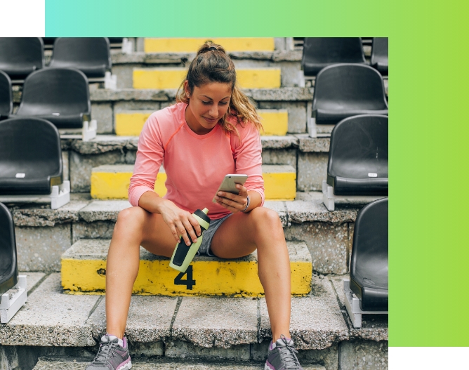 A women sits on the steps of a stadium holding a water bottle and looking at her smartphone.