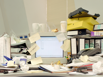 Image of an office desk cluttered with notepads, binders, paper, sticky notes, and used coffee cups