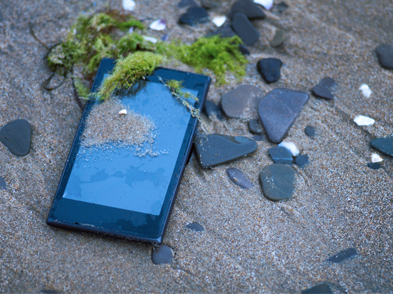 Cell phone laying on the beach covered with sand, rocks and seaweed