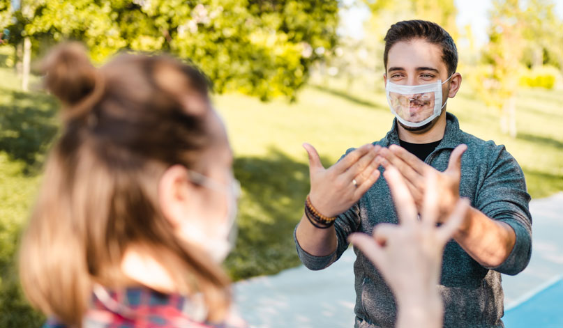 Man wearing a clear see through mask uses sign language to communicate with woman.