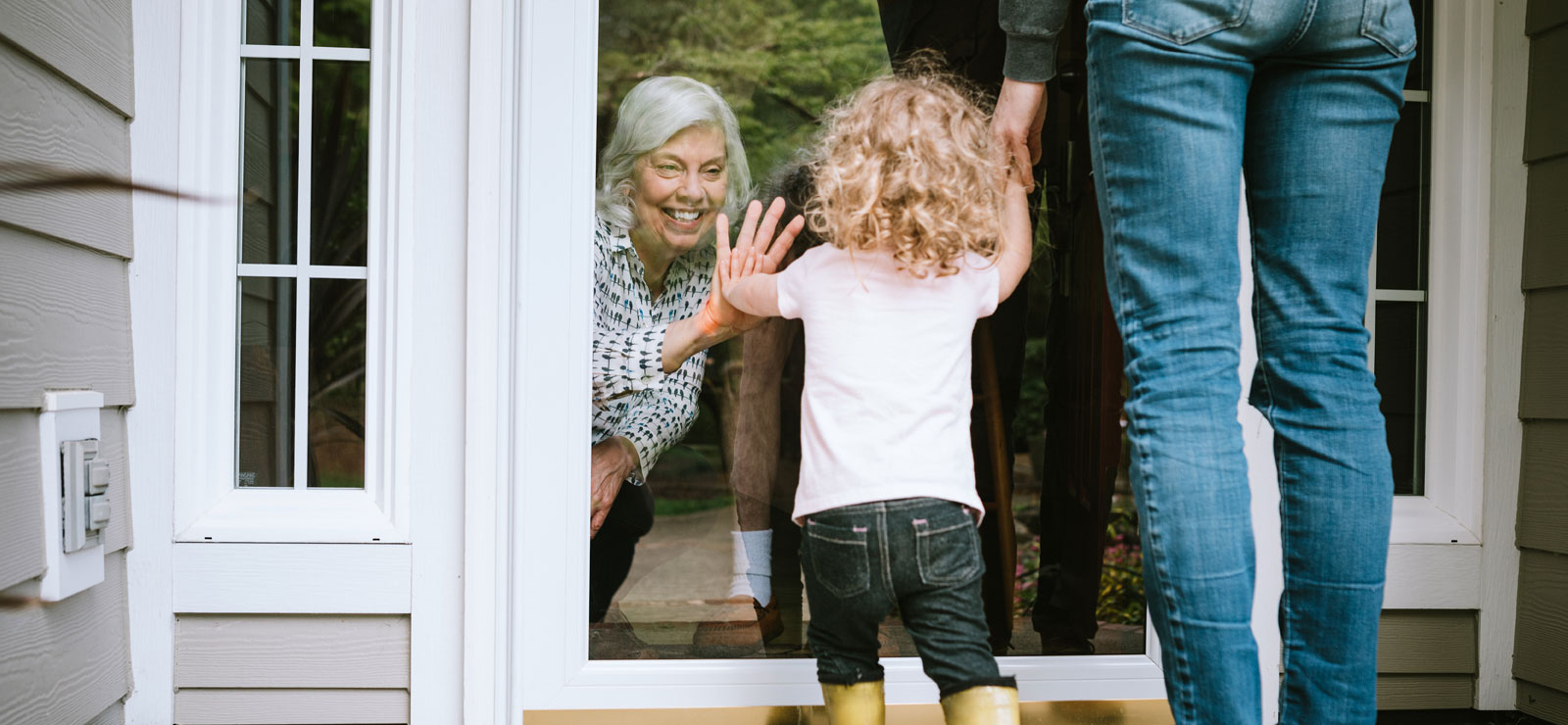 Grandma standing behind glass door puts hand against door while granddaughter on the other side of the door does the same thing.