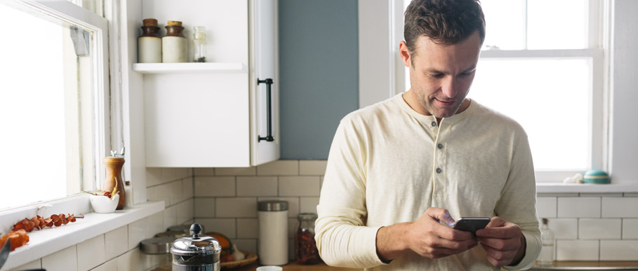 A man stands in his kitchen typing into his smartphone