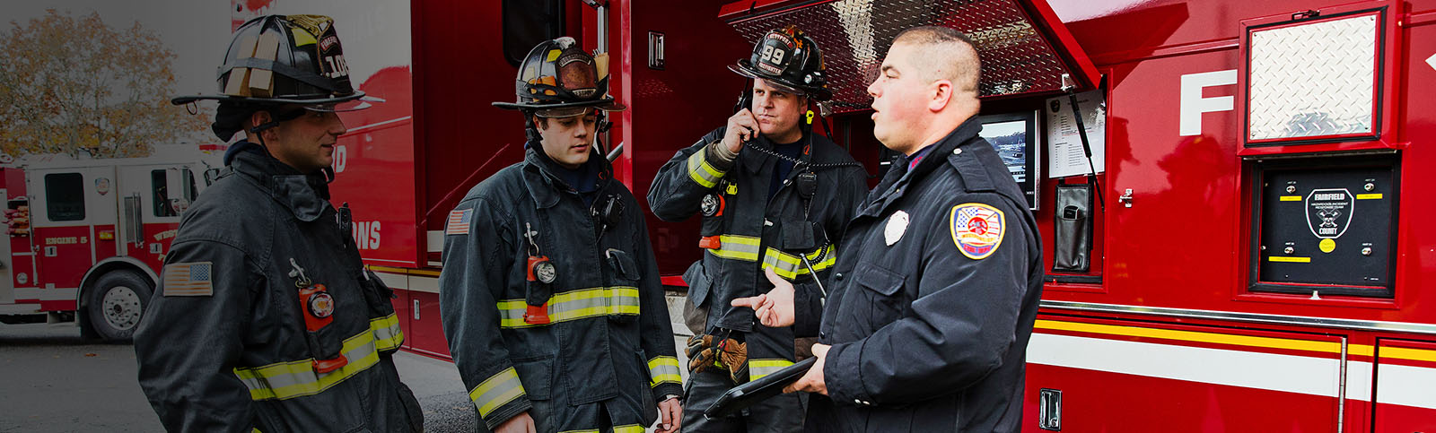 Four firefighters in uniform stand and talk by a firetruck.  