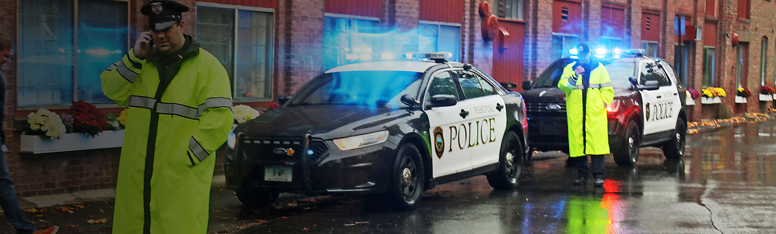 Police officers standing in the rain in bright yellow reflective rain coats and police hats standing in front of two police cars with lights on, one using a cell phone while the other appears to look at phone screen  