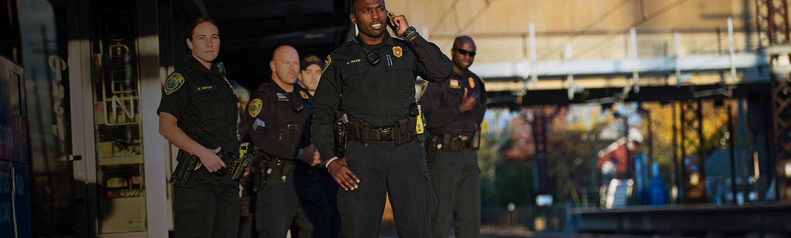 Group of police officers in black uniforms standing next to a train platform as they appear to await for the train, one of whom is talking on the phone 