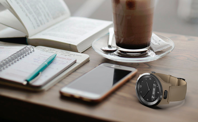 The Navigil 580 wellness wristwatch sits on a desk next to a smartphone, a planner, a book, and a cup of ice coffee