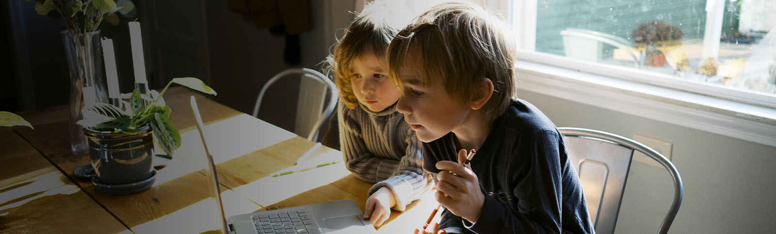 Kids in morning light sitting at table doing homework on laptop.
