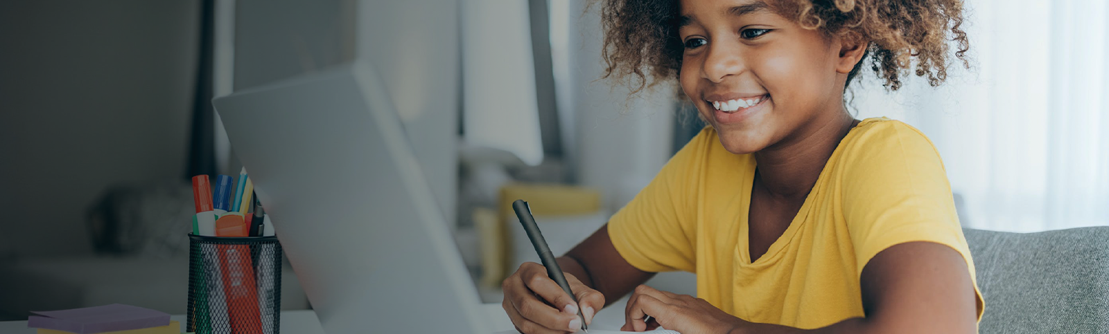 Elementary-aged child smiling while holding a pen doing school work while sitting in front of a computer.