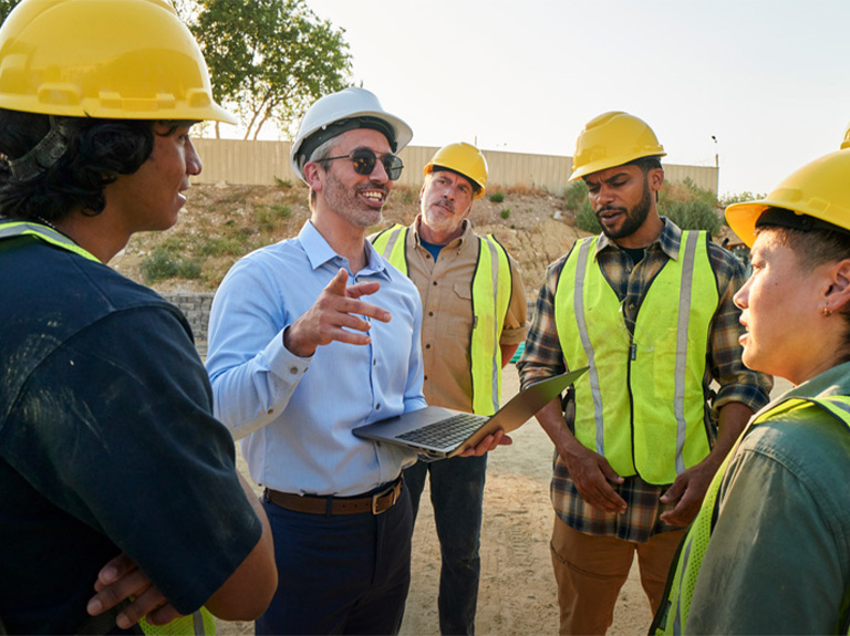 Man standing holding laptop outside smiling, surrounded with other people, all wearing safety helmets. 