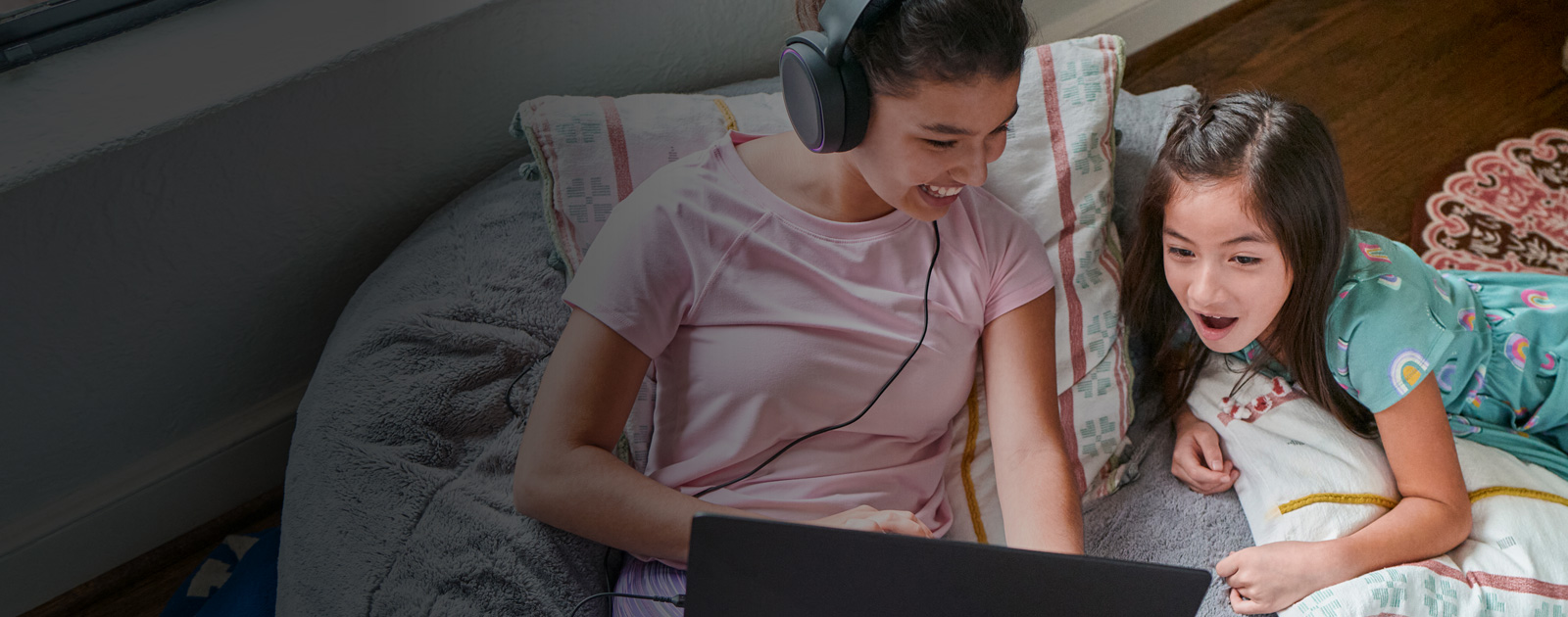 Older sister smiling wearing headphones, showing younger sister laptop.