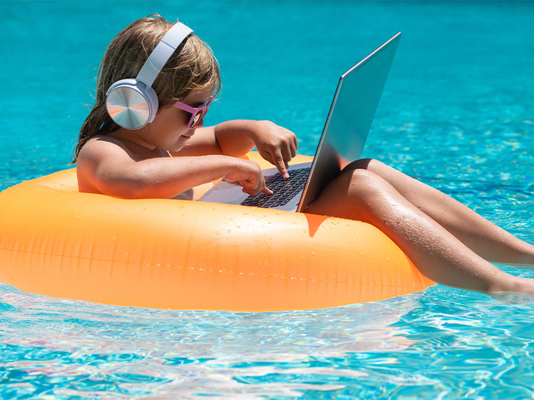 Kid online working on laptop, swimming in a sunny turquoise water pool.